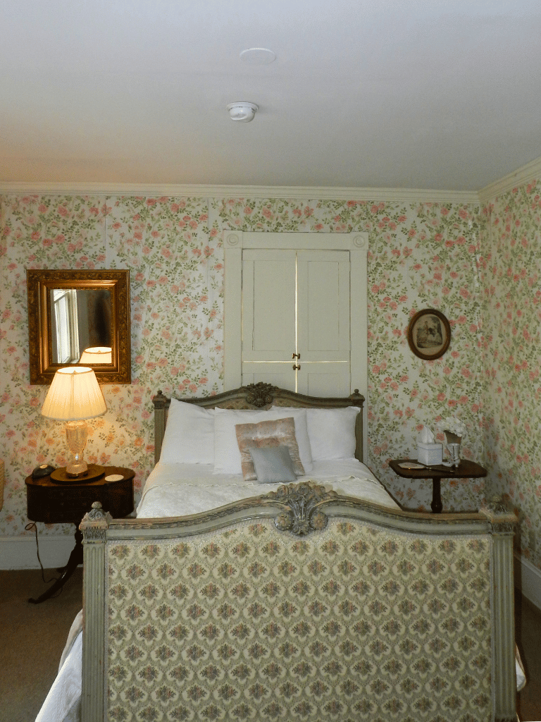 Double bed with a floral patterned and wooden bedframe in the Monroe Room (Room 24) at The Historic Wayside Inn 1797, featuring a private full bathroom and period antiques in Middletown, VA.